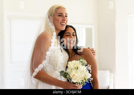 Embracing bridesmaid, bride holding bouquet, both smiling joyfully in bright room Stock Photo