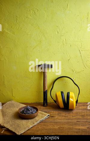 Still life with construction tools: hammer, nails in a wooden bowl, and protective earmuffs on a yellow textured wall background. Concept of building, Stock Photo