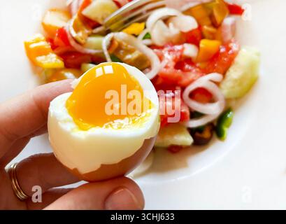 Hand holding a soft-boiled egg, fresh salad in a plate on a white background. Healthy food. Focus on the foreground. Close-up. Stock Photo