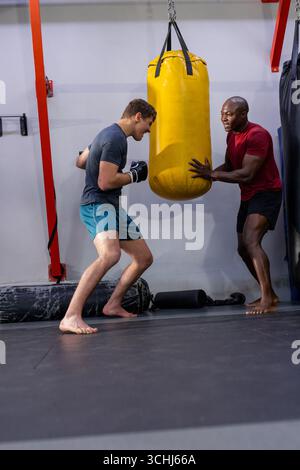 African american male coach with male trainee in sportswear practicing punches on yellow bag in gym Stock Photo