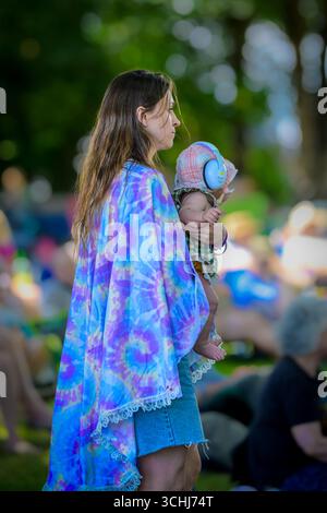 Baby with hearing protectors held by woman  wearing tie dyed shawl at Music Festival Stock Photo