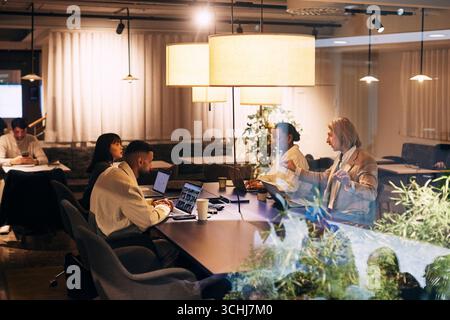 Male and female business colleagues discussing ideas with each other while sitting at desk in illuminated open plan offi Stock Photo