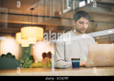 Focused male business entrepreneur working on laptop while sitting at desk in coworking office seen through glass Stock Photo
