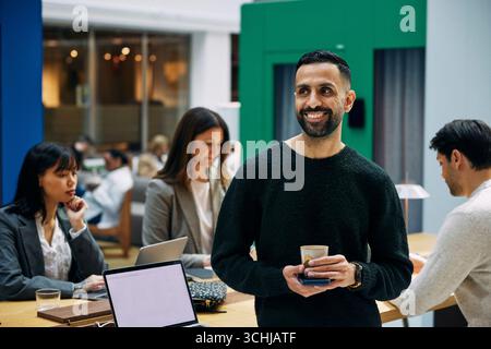 Smiling male entrepreneur holding mug looking away while standing near table in office Stock Photo
