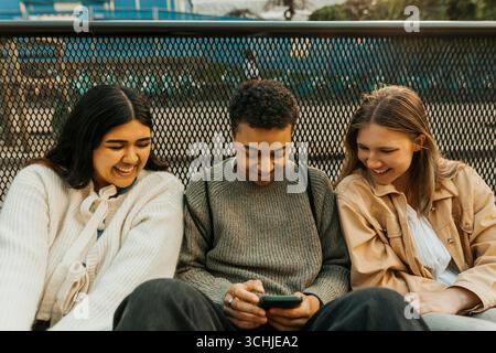 Happy young women sitting with male friend using smart phone near railing Stock Photo