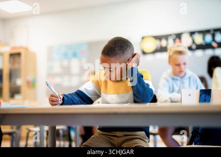 Boy writing in paper while sitting at desk in elementary school classroom Stock Photo
