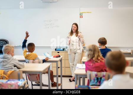 Female teacher teaching mathematics to group of elementary students sitting at desk in classroom Stock Photo