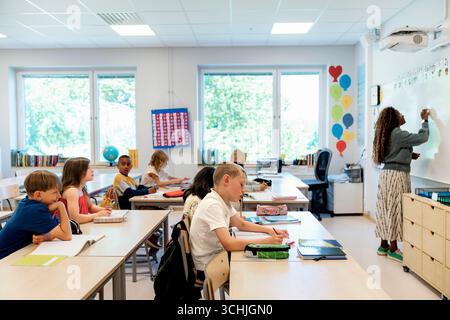 Group of elementary students studying sitting at desk while teacher writing on whiteboard in classroom Stock Photo