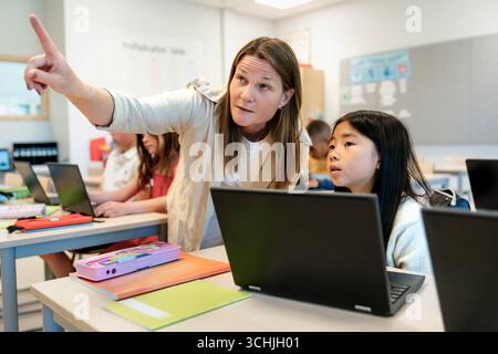 Female teacher pointing while standing next to girl sitting at desk in elementary classroom Stock Photo