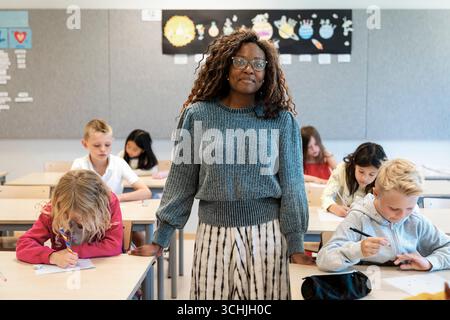 Portrait of smiling female teacher standing amidst students sitting at desks in classroom Stock Photo