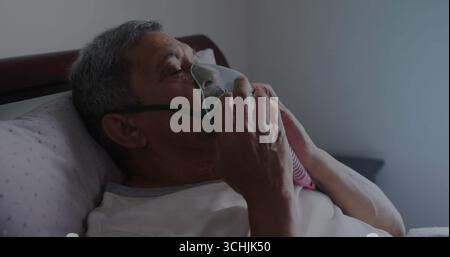 Holding oxygen mask tubing man wearing white tee lying on bed in bedroom, with pillow, nightstand Stock Photo