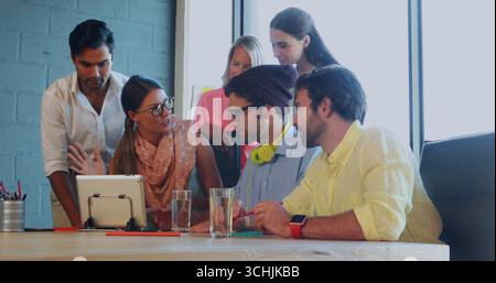 Collaborating six office professionals leaning over table in meeting room with tablet on stand Stock Photo