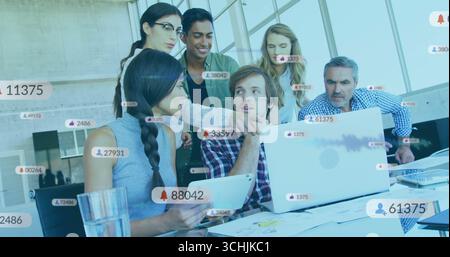 Analyzing team of six working on laptops and tablet at modern office, with floating media icons Stock Photo