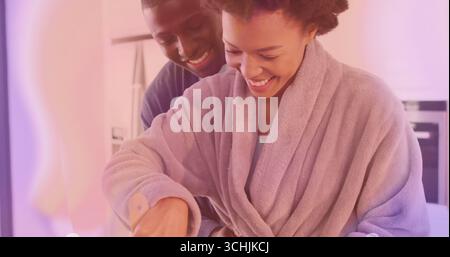 Bathrobe wearing woman cooking with smiling partner in kitchen, with wooden spatula and mixing bowl Stock Photo