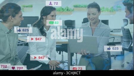 Collaborating colleagues analyzing social media icons on laptop in open-plan office, with tablet Stock Photo