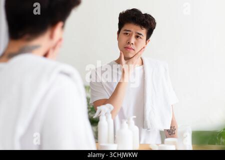Cheerless asian young man with tattoos in white t-shirt, with towel, touches hand to cheek, checks stubble Stock Photo