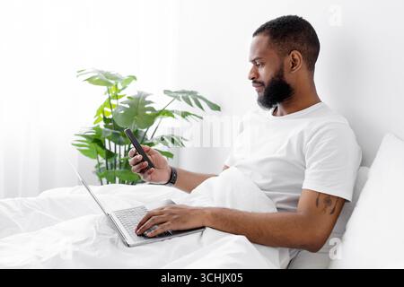 Serious millennial african american bearded guy sit on bed in bedroom interior, working on laptop Stock Photo