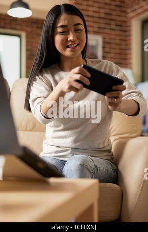 Positive asian entrepreneur seated in living room, grasping smartphone with both hands, immersed in online video game. Close up of smiling businesswoman on joyful break from work duties. Stock Photo