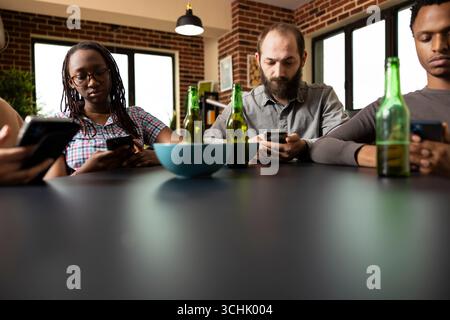 Mixed group of young adults using smartphones, casually sitting with snacks and drinks, focused on social media while hanging out. Group of friends browsing or texting on their mobile devices. Stock Photo