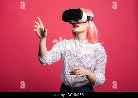 Young woman wearing pink wig and VR headset, lifting hand as she interacts with advanced 3D environment. Caucasian lady with virtual reality glasses standing and exploring futuristic digital world. Stock Photo