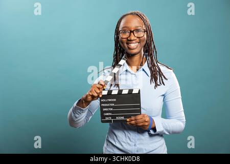 Cheerful female filmmaker looks at camera, holding a clapperboard, standing isolated over a blue background. Happy african american woman with a film slate in hand, excited of new movie project Stock Photo