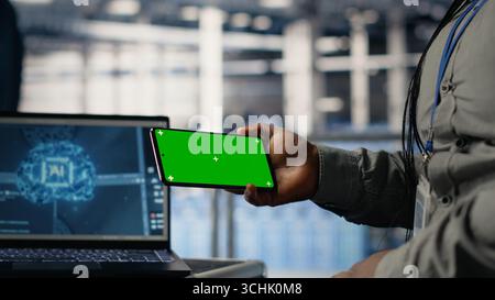 Data center worker uses green screen smartphone while deploying deep learning software. Server hub employee uses mockup mobile phone while training AI systems with datasets Stock Photo