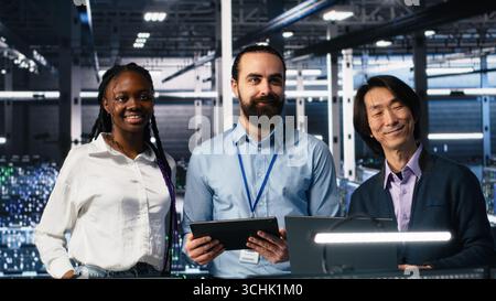 Portrait of smiling engineers in server room using AI to review infrastructure diagnostics. Cheerful IT coworkers in data center analyzing performance data using artificial intelligence, camera A Stock Photo