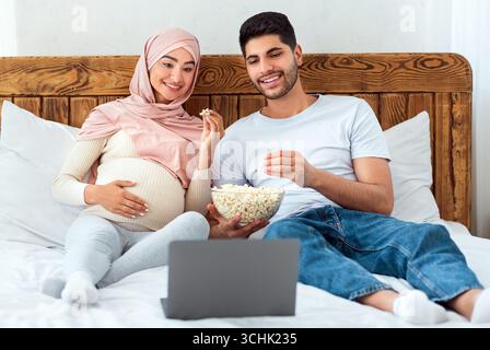 Leisure time. Relaxed arab man and pregnant wife watching film on laptop computer, eating popcorn, sitting on bed Stock Photo