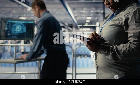 Data center programmers check AI dashboard using tablet, doing systems review. Server hub coworkers test artificial intelligence tools on device to ensure reliable performance, camera A Stock Photo