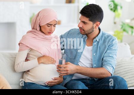 Care and support during pregnancy. Muslim expecting woman feeling sick, man giving her glass of water, siting on sofa Stock Photo