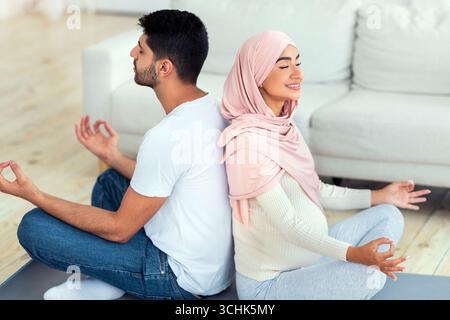 Yoga for pregnancy. Happy muslim spouses meditating together at home, sitting back to back and practicing lotus pose Stock Photo
