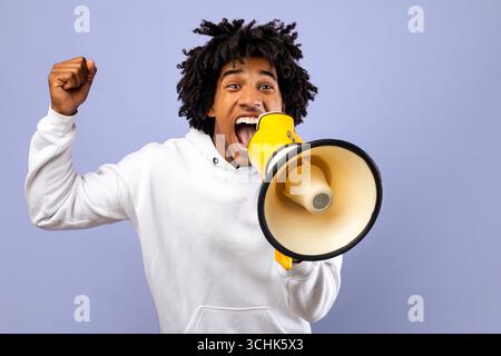 Agitated black teenager shouting into megaphone over violet studio background Stock Photo