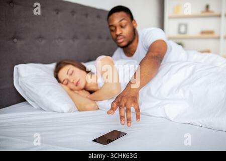 Suspicious black husband checking his sleeping wife's cellphone in bed at home. Relationship problems Stock Photo