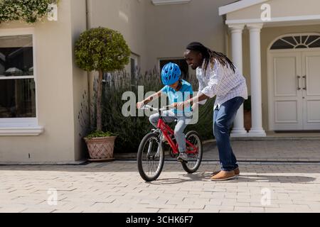 Father teaching son to ride bicycle in front of house, both smiling Stock Photo