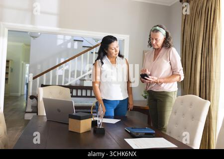Two women discussing smartphone features at home office with laptop and documents Stock Photo