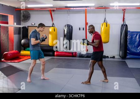 Diverse male trainer with trainee practicing sparring in martial arts gym with gloves and mitts Stock Photo