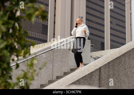 Woman descending stairs at office talking on smartphone, coffee cup, crossbody bag, copy space Stock Photo
