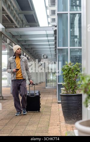African american man pulling suitcase while using smartphone on walkway near planters Stock Photo