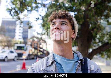 Man looking upward on city sidewalk wearing denim jacket, knit beanie, backpack Stock Photo