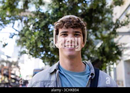 Man standing on city sidewalk wearing beige beanie, denim jacket, teal shirt, holding backpack Stock Photo