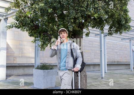 Man calling on smartphone while holding black rolling suitcase, backpack on urban plaza Stock Photo