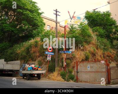 art school sign in Enna Sicily with rustic truck full of boxes under old electricity pole and lush greenery on hillside urban street corner Stock Photo