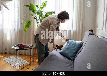 Serene woman arranging pillows on sofa while organizing living room in tidy comfortable home Stock Photo