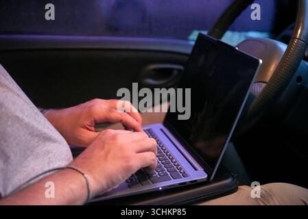 Unrecognizable person using a laptop sitting in a car, it is nighttime and raining outside. Concepts: digital nomad, remote work and working while tra Stock Photo
