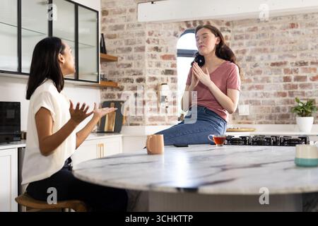 Engaging in lively conversation, two young women sit in modern kitchen Stock Photo