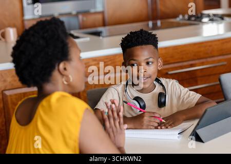 Mother helping son with homework at kitchen table, using tablet and notebook Stock Photo