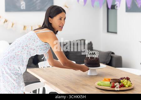 Setting chocolate birthday cake on wooden table, Asian woman in floral dress smiles Stock Photo