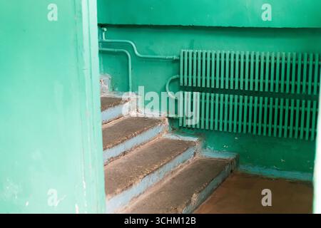 Interior of an old building in poor condition with green painted walls, cracked plaster, radiator pipes, handrail, and worn flooring. Urban decay Stock Photo