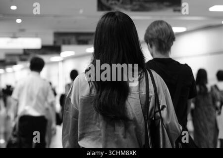 Back view of woman in urban corridor surrounded by crowds, evoking introspection, movement, and quiet focus within the anonymity of city life. Stock Photo