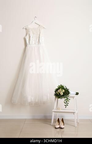 Beautiful wedding dress hanging on white wall, wedding shoes and bridal bouquet in room Stock Photo